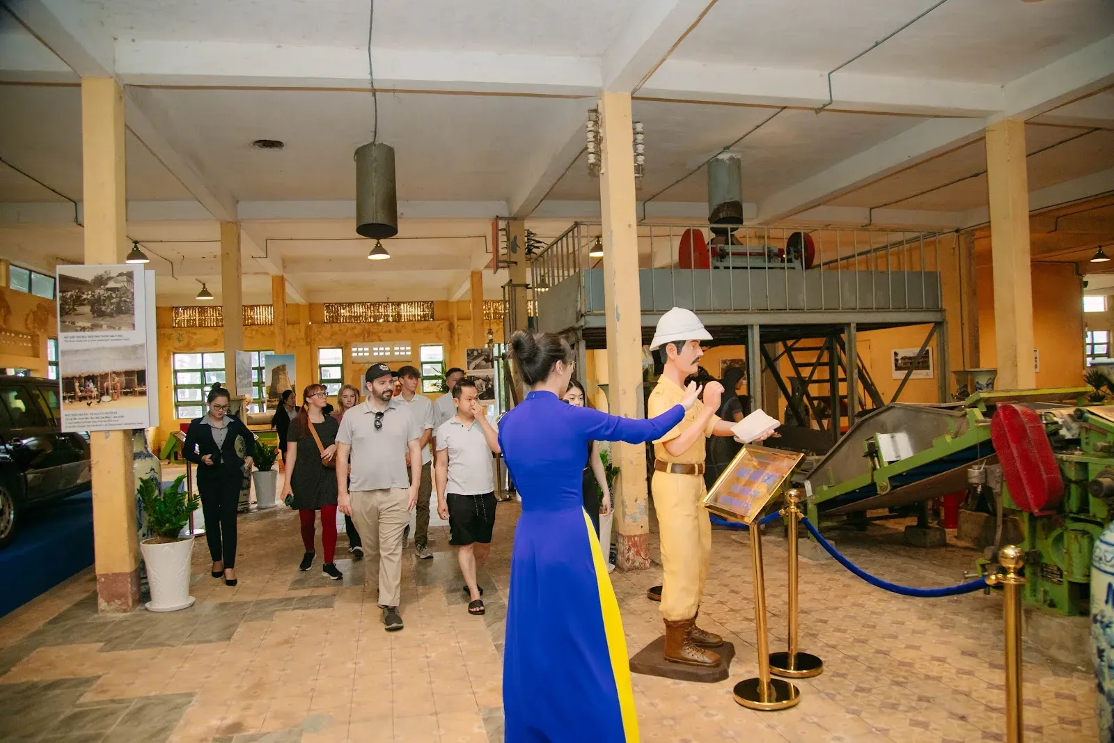 Thuyết minh viên áo dài xanh giới thiệu với đoàn khách bên tượng người công nhân trong xưởng